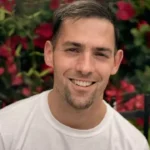 Uśmiechnięty mężczyzna z lekkim zarostem sfotografowany na tle czerwonych kwiatów